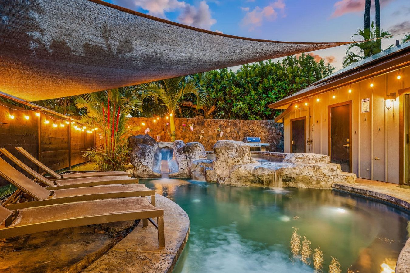 Une piscine avec une cascade, des chaises longues, un barbecue et un mur en pierre.