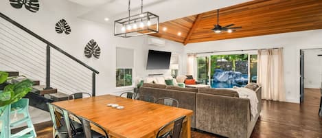 A spacious living area with a wooden ceiling, a dining table with chairs, a sofa, and a television.