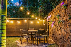 Een terras buiten met een tafel, stoelen, lichtslingers, een bamboepilaar en een stenen muur.