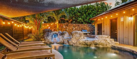 A pool with a waterfall, lounge chairs, a grill, and a stone wall.