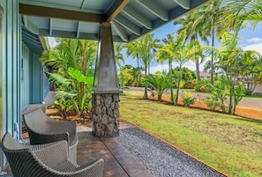 A covered outdoor seating area with wicker chairs, a stone pillar, and a view of a well-maintained lawn with palm trees.