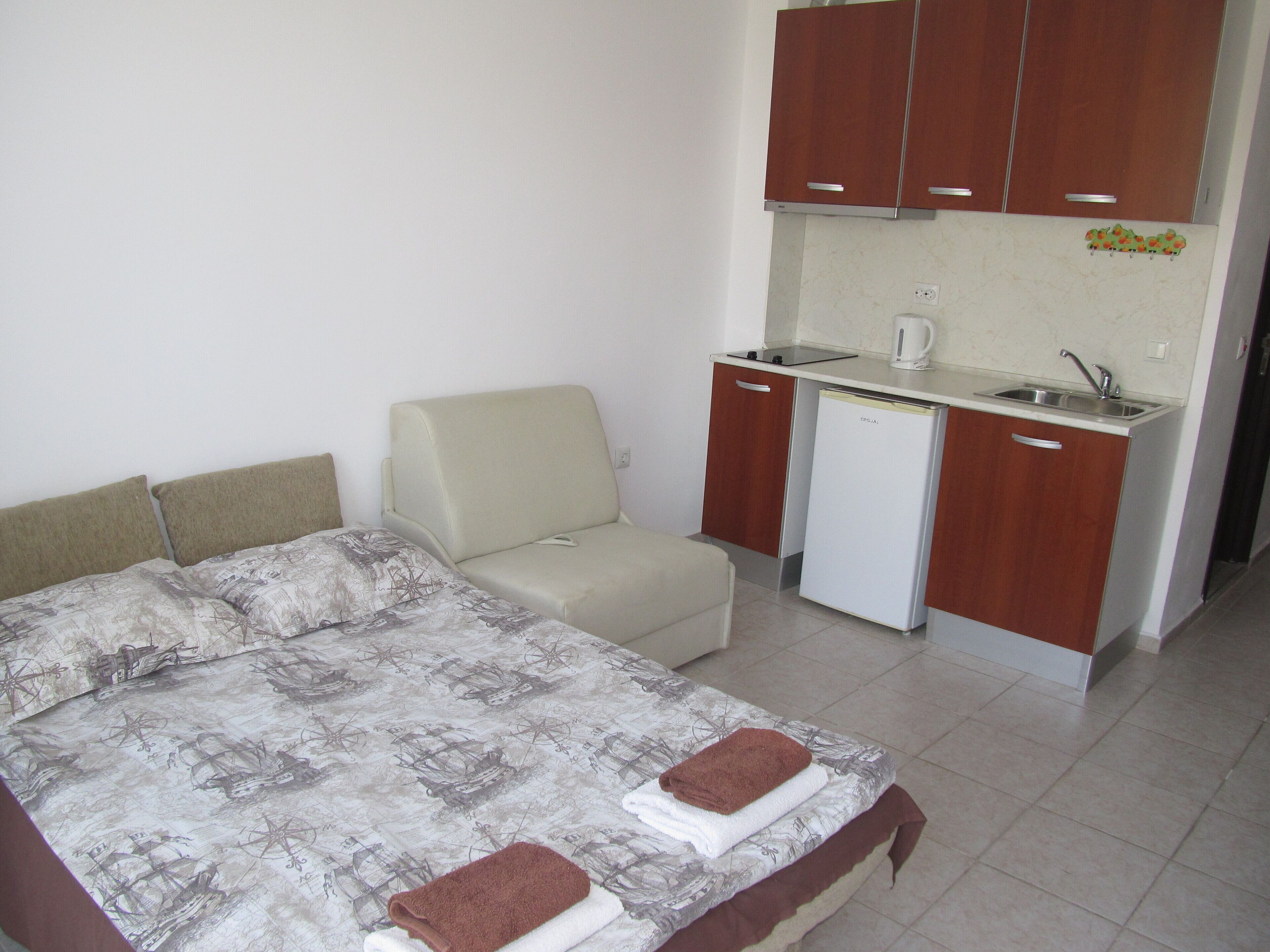 A compact room with a bed, a small sofa, a kitchenette, and a compact refrigerator.
