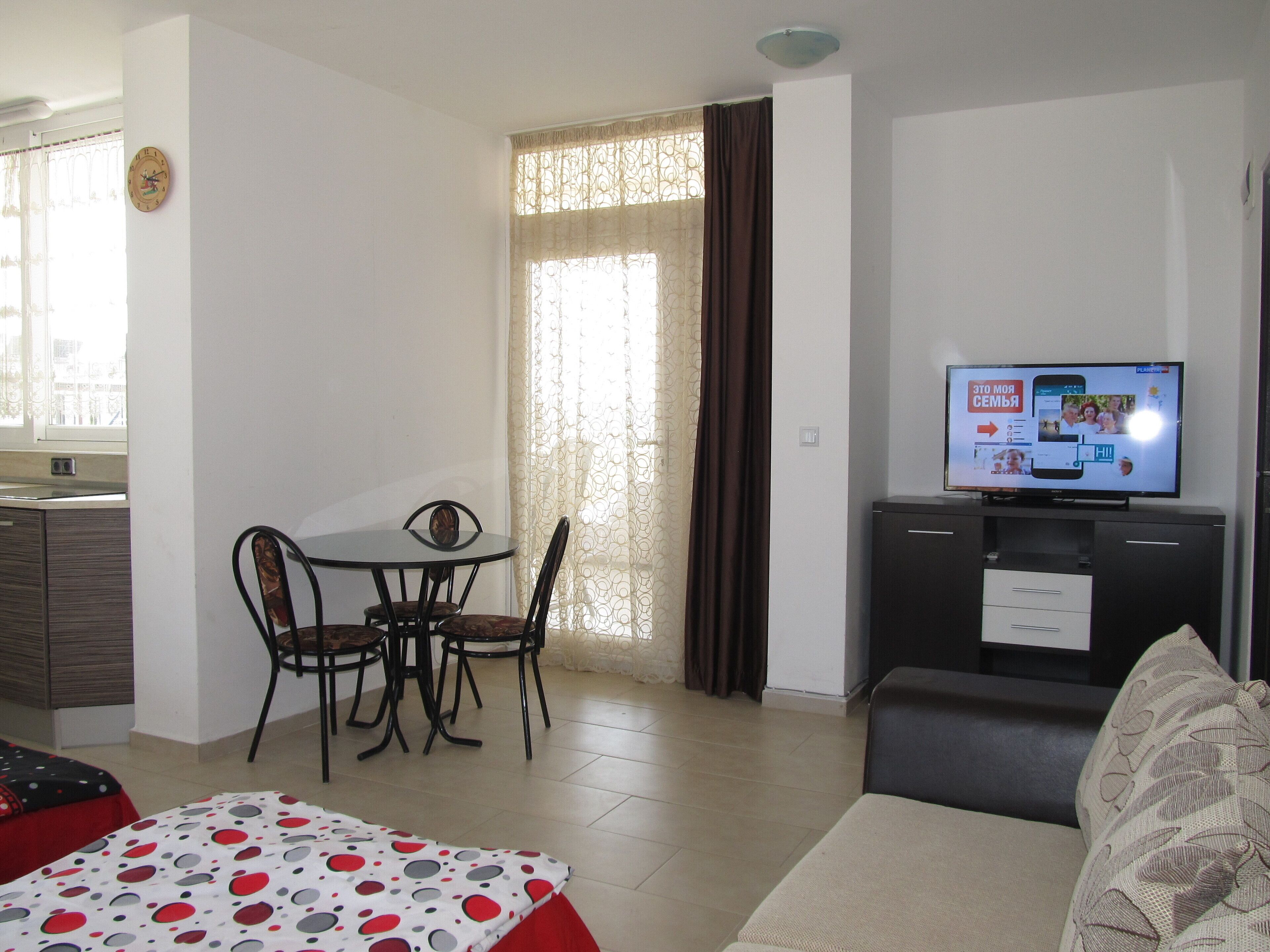 A living room with a sofa, a TV, a dining table with chairs, and a bed with a patterned bedspread.