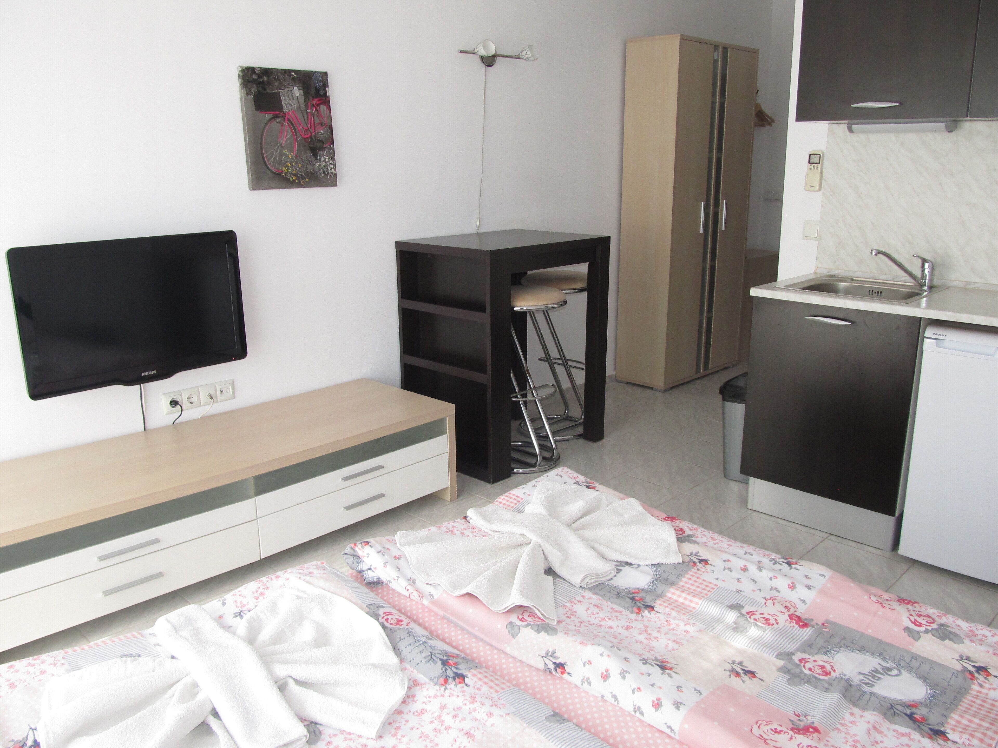 A compact room with a flat-screen TV, a wooden cabinet, a small kitchen area with a sink, and a bed with a floral-patterned bedspread.