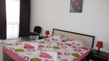 Un lit bien fait, recouvert d’une couvre-lit à motifs, une chaise noire, une lampe rouge et un tableau encadré accroché au mur.