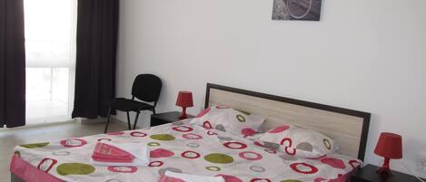 A neatly made bed with a patterned bedspread, a black chair, a red lamp, and a framed picture on the wall.