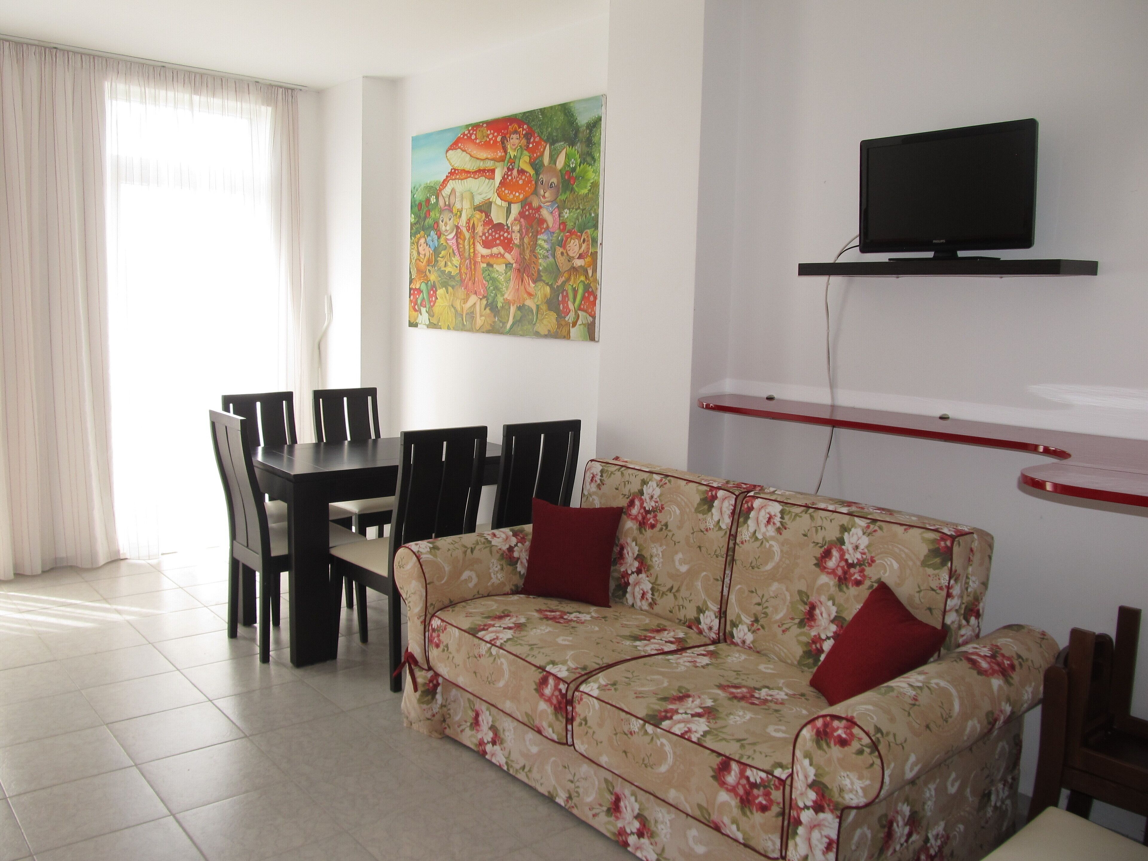 A living room with a floral sofa, a dining table with chairs, a flat-screen TV mounted on a red shelf, and a colorful painting on the wall.