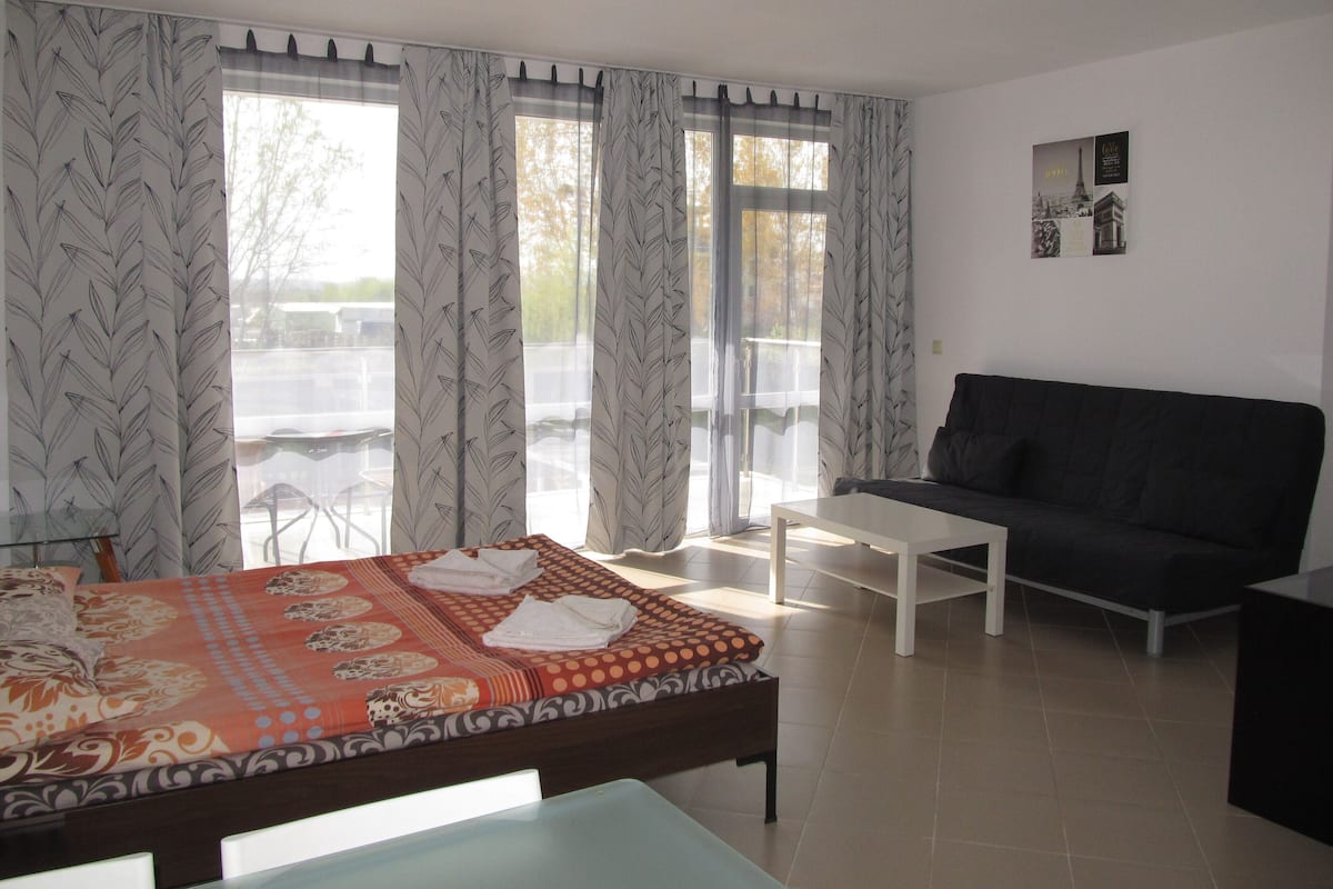 A hotel room with a bed, a sofa, a small table, and large windows with curtains.