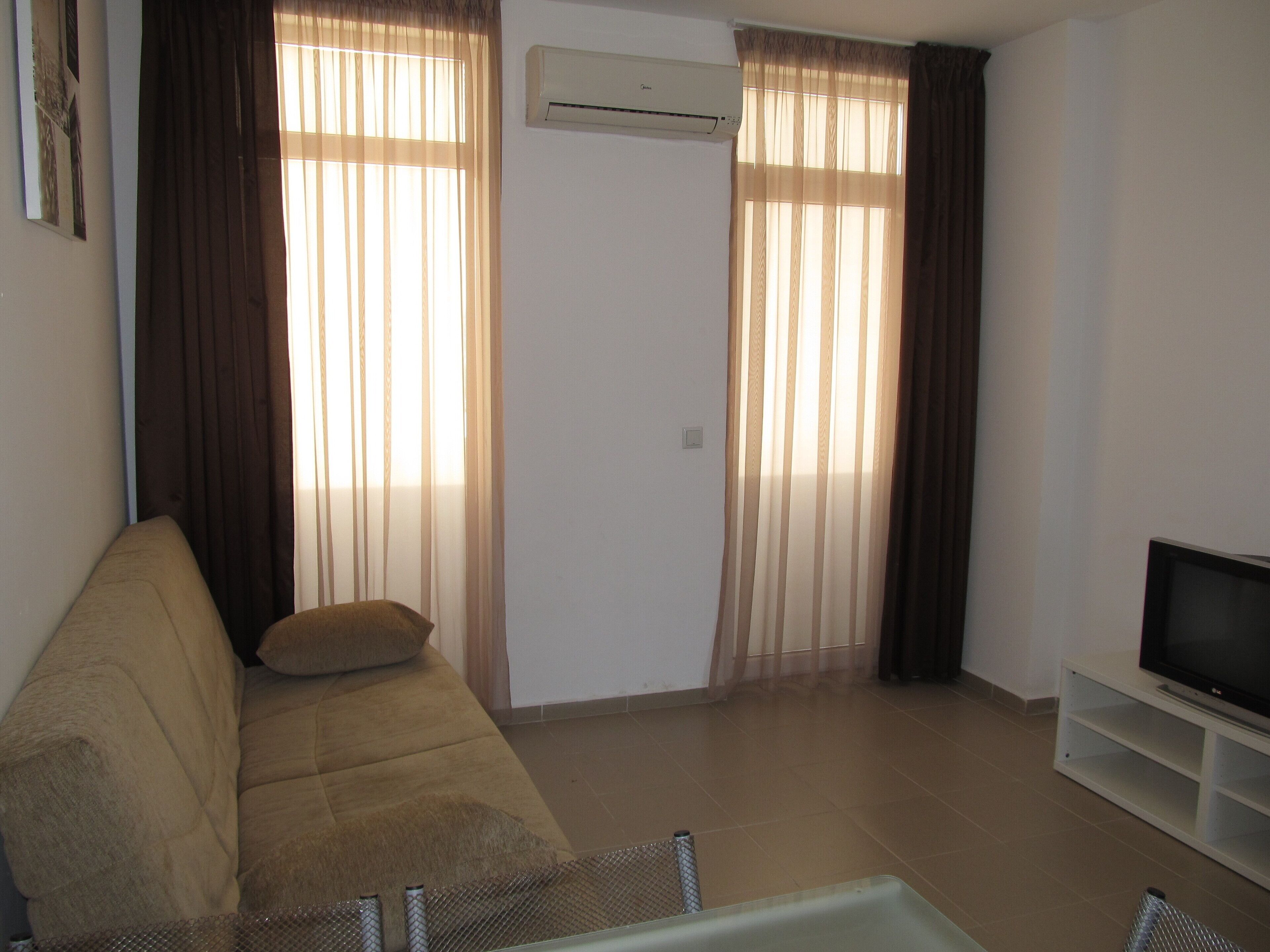 A living room with a beige sofa, a TV on a stand, and a sliding glass door with curtains.