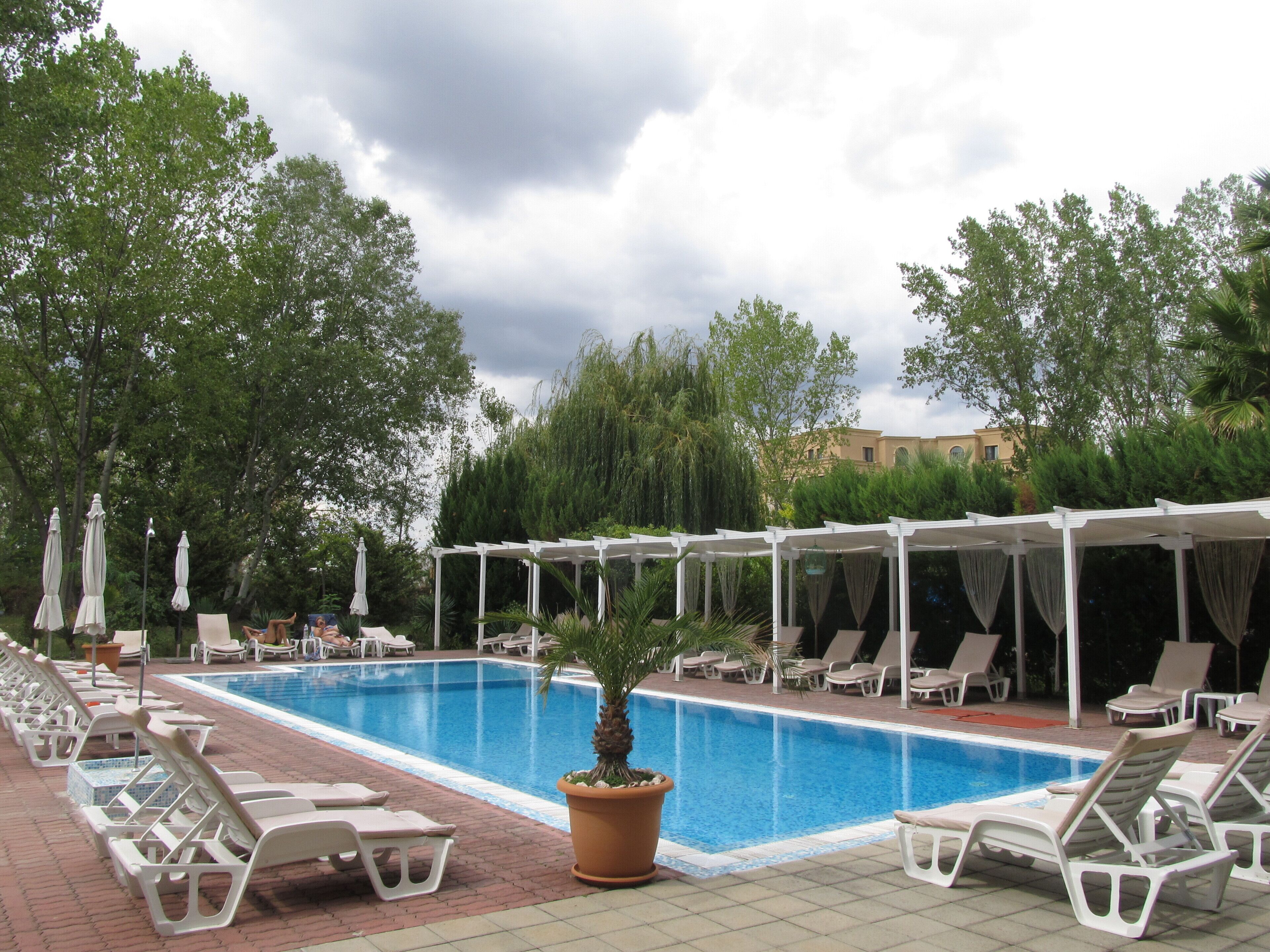 Una piscina con sillas de descanso y sombrillas, rodeada de árboles y un edificio al fondo.
