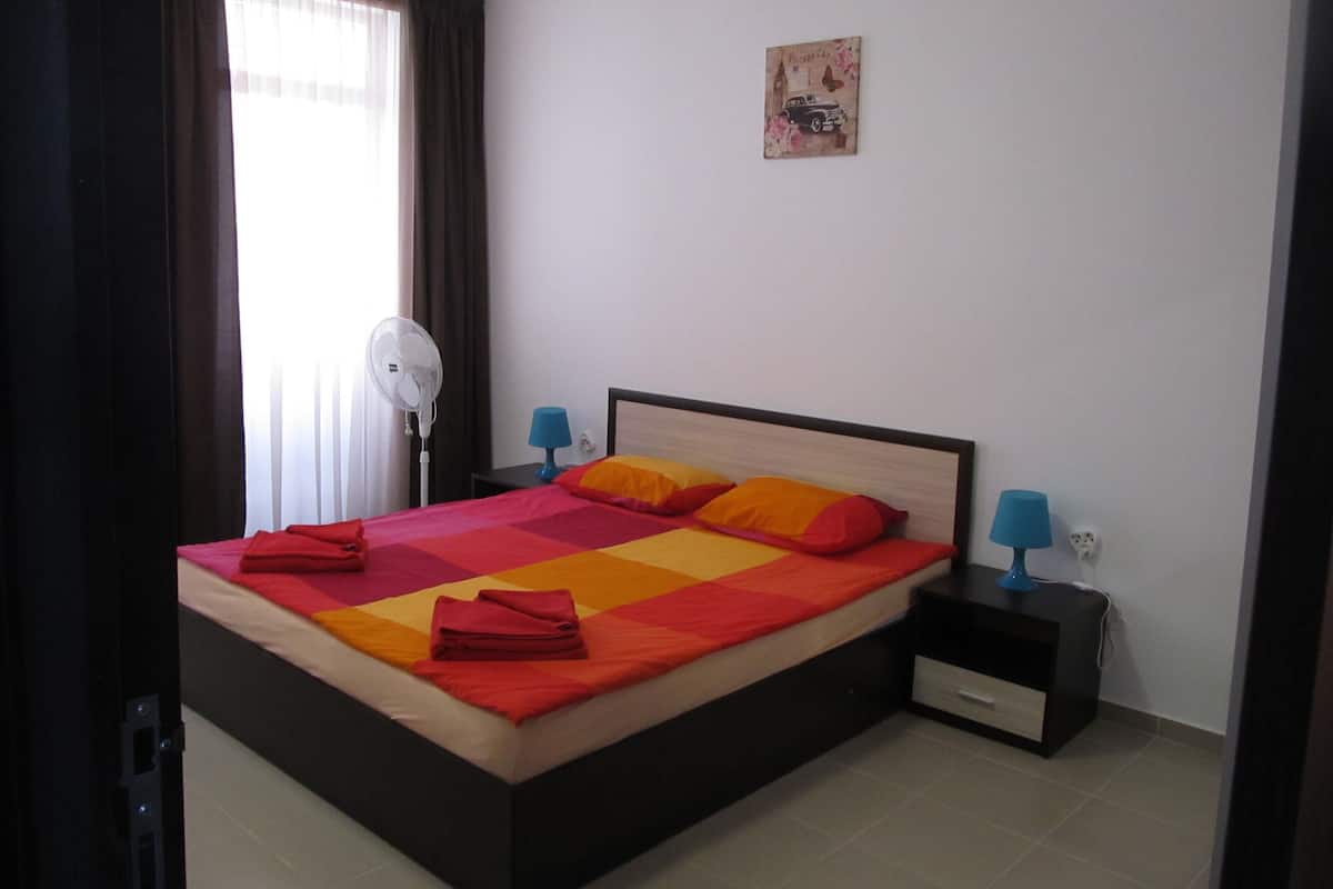A bedroom with a bed, two bedside tables, a wall-mounted fan, and a small picture.