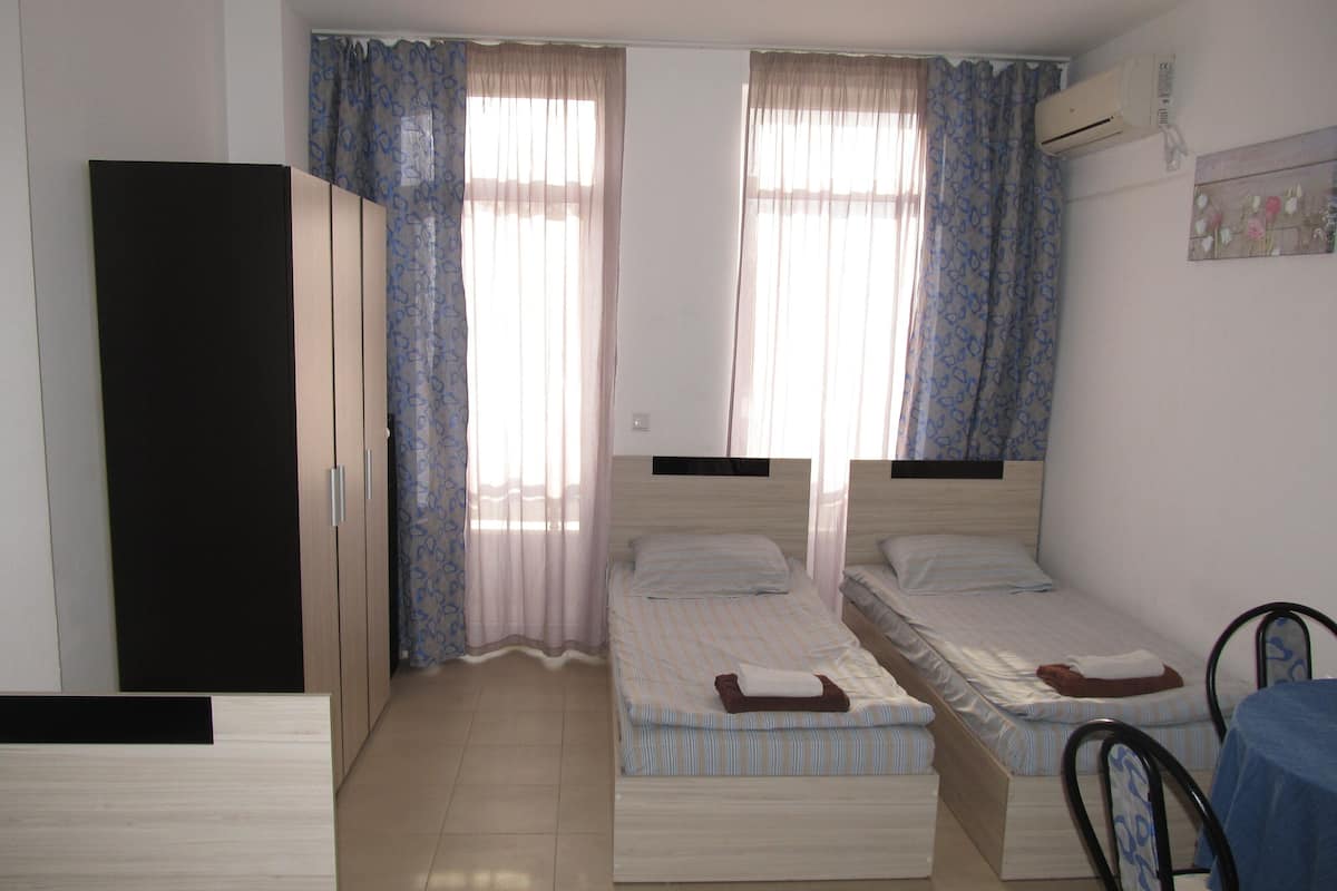 A bedroom with two single beds, a wardrobe, a table with a chair, and an air conditioning unit.