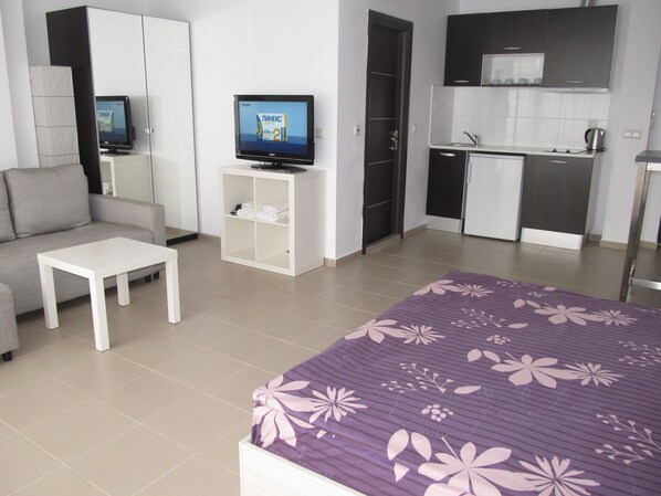 Une chambre d’hôtel moderne avec un canapé, une petite table blanche, un lit recouvert d’une courtepointe à motifs floraux violets, un téléviseur posé sur un meuble et une kitchenette équipée d’un évier et de placards.