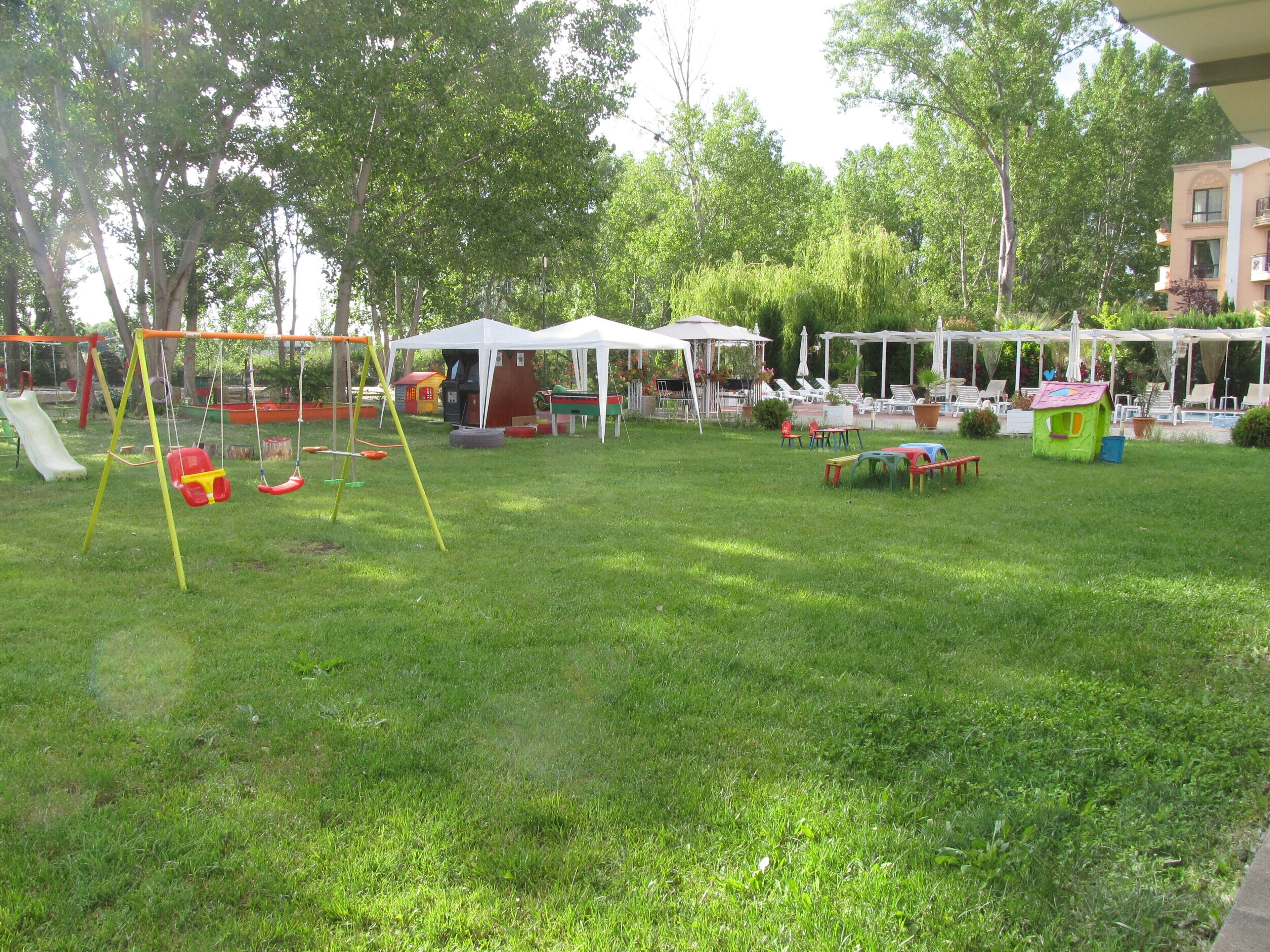 Ein grünes Außenbereich mit Schaukeln, Picknicktischen und einem kleinen Spielhäuschen.
