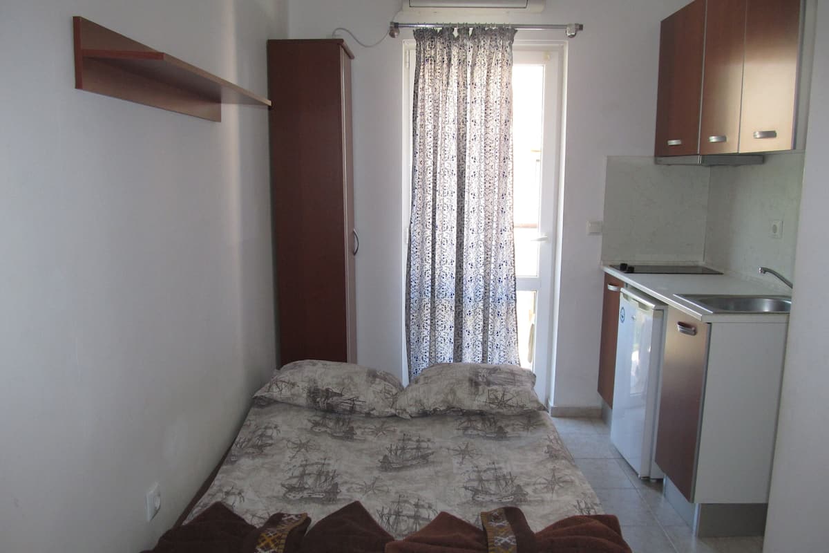 A small, compact room with a bed, a kitchenette, and a window with curtains.