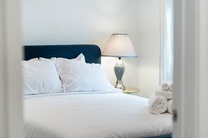 Une chambre d’hôtel avec un lit, du linge de lit blanc, une tête de lit bleue, une lampe de chevet et une fenêtre avec des serviettes roulées.