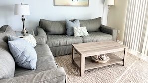Una sala de estar con un sofá gris, una mesa de centro de madera y un cuadro con motivos playeros en la pared.