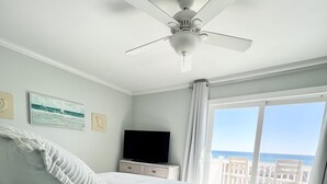 Un dormitorio con una cama grande, un ventilador de techo, un televisor sobre un mueble de madera y vistas al mar a través de amplios ventanales.