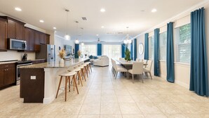 Une cuisine moderne dotée d’un îlot central, de tabourets de bar, d’un coin repas avec une table et des chaises, et d’un salon avec un canapé et un téléviseur.