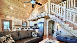 A spacious living area with a leather sofa, a coffee table with magazines, a wooden staircase, and a kitchen in the background. - Luxury Mountaintop Getaway with, Fire Pit, New Hot Tub, WiFi & Gorgeous Views! (Sevierville)