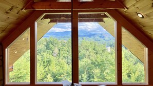 A room with a sloped ceiling and large windows offering a view of a forested mountain range. - Luxury Mountaintop Getaway with, Fire Pit, New Hot Tub, WiFi & Gorgeous Views! (Sevierville)