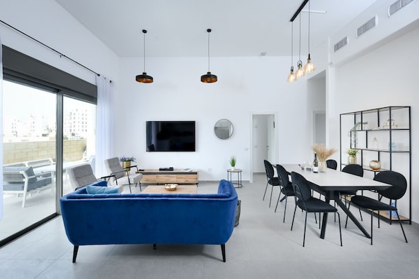 Ein modernes Wohnzimmer mit einem blauen Sofa, einem Essbereich mit weißem Tisch und einem an der Wand befestigten Fernseher.