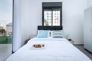 Ein modernes Schlafzimmer mit einem großen Bett, einem Fenster mit Blick auf Gebäude, einem Nachttisch mit Lampe und einer Schale mit Gebäck.