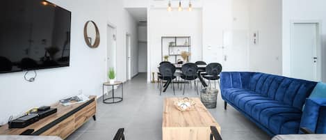 Een moderne woonkamer met een grote televisie, een blauwe bank, een houten salontafel, en een eethoek met stoelen en een tafel.