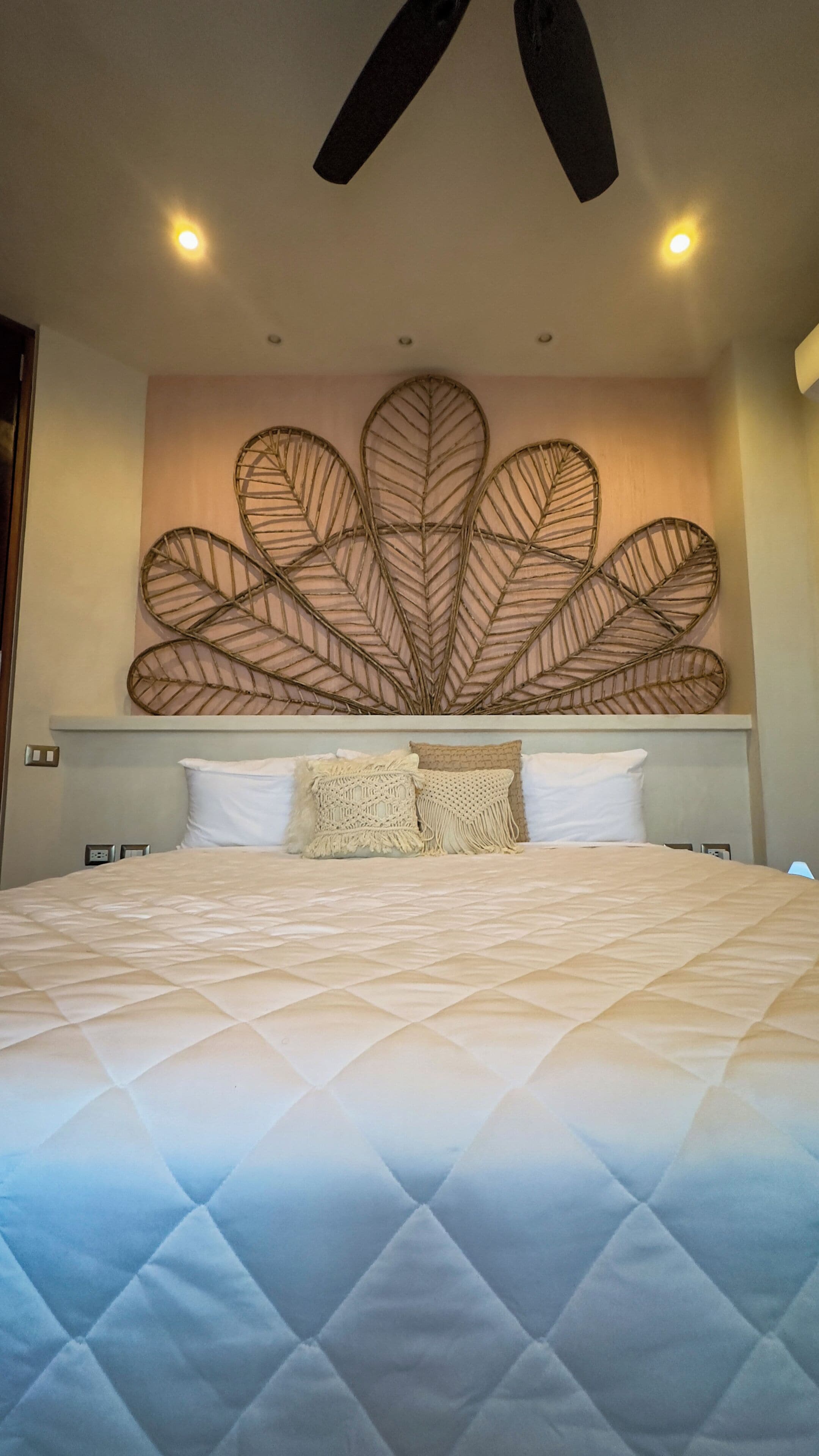 A bed with a quilted duvet, a wall-mounted leaf-patterned decor, and a ceiling fan.