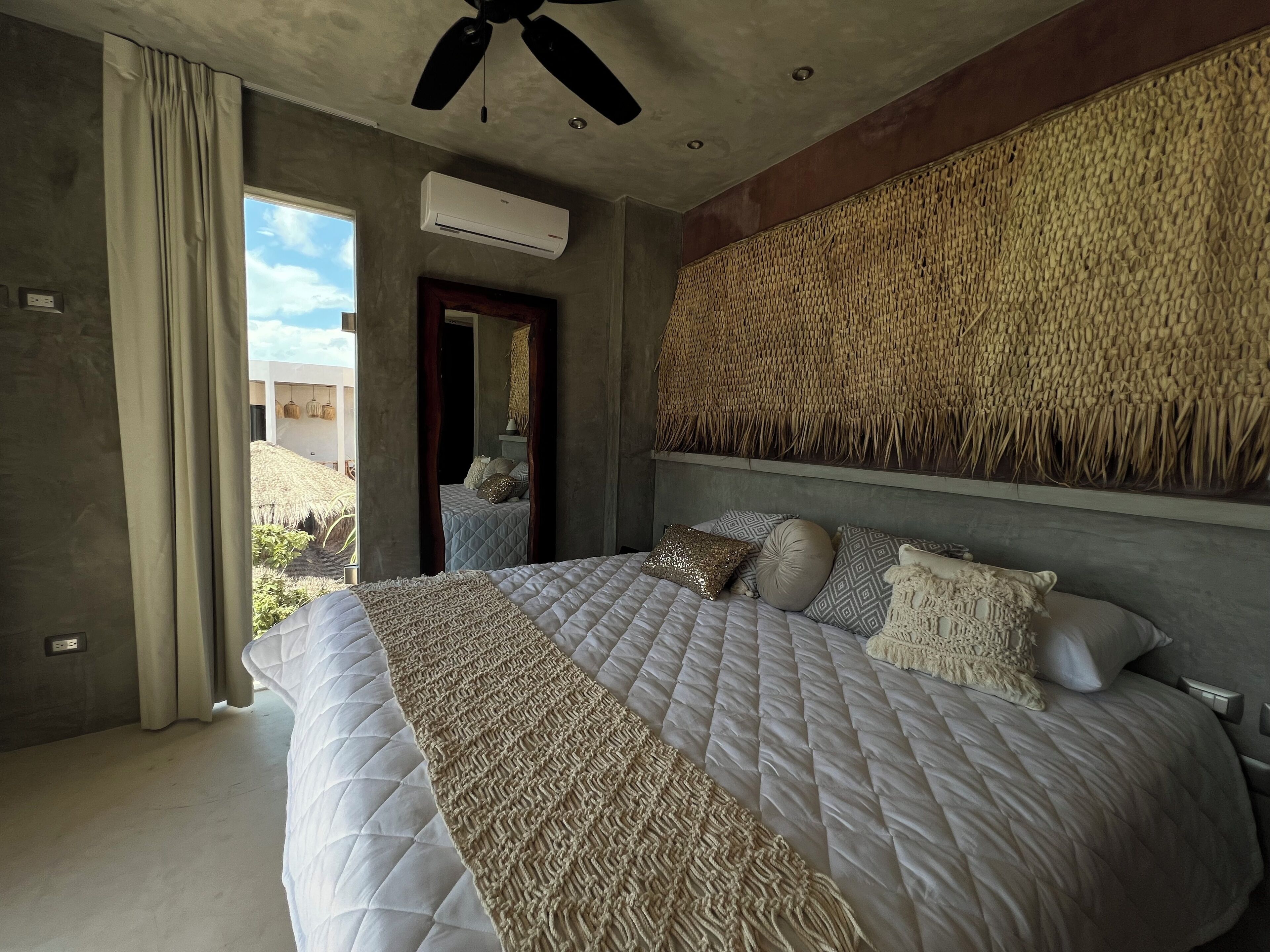 A bedroom with a large bed, a ceiling fan, an air conditioning unit, and a woven wall hanging.