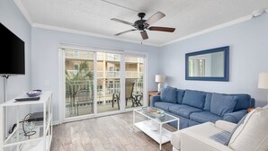 A living room with a blue sofa, a white armchair, a coffee table, a TV, and a ceiling fan. - St. Simons Beach Club, Oceanfront Pool, Fabulous Beach Access and More! (Saint Simons Island)