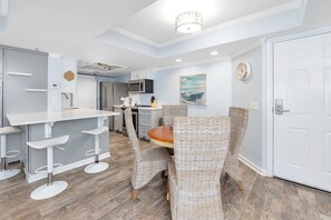 A modern kitchen with white cabinets, a microwave, and a coffee machine. There is a dining area with wicker chairs and a wooden table.