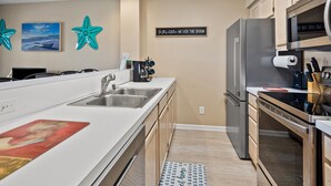 Una cocina moderna con un lavamanos de acero inoxidable, horno empotrado y refrigerador. Hay estrellas de mar decorativas en la pared y un cartel que dice 'En la cocina, lamemos la cuchara'.