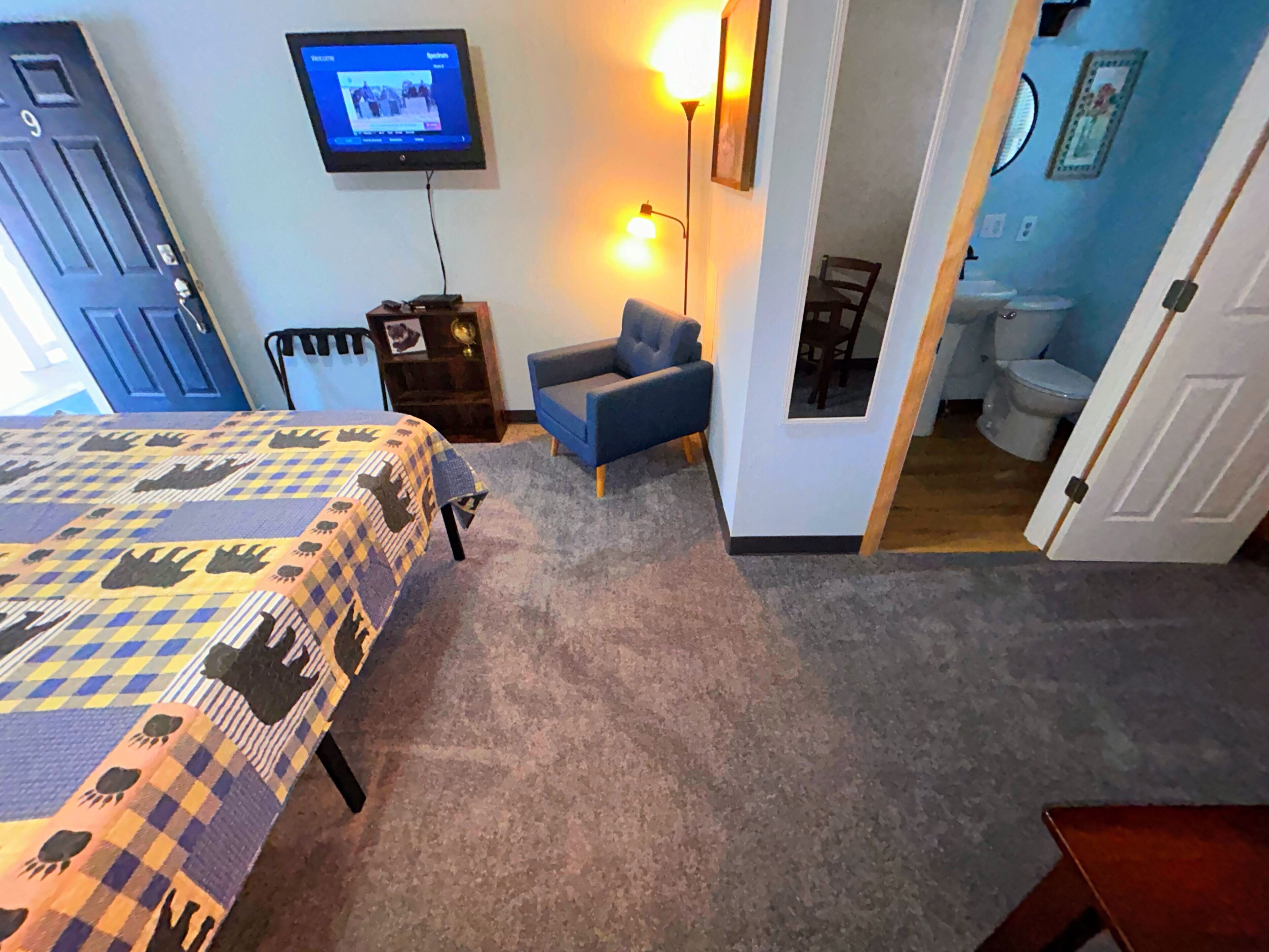 A bedroom with a bed, a TV, a chair, and a bathroom visible through an open door.