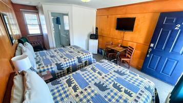 A hotel room with two beds, a small table, a chair, a TV, and a door with a number.