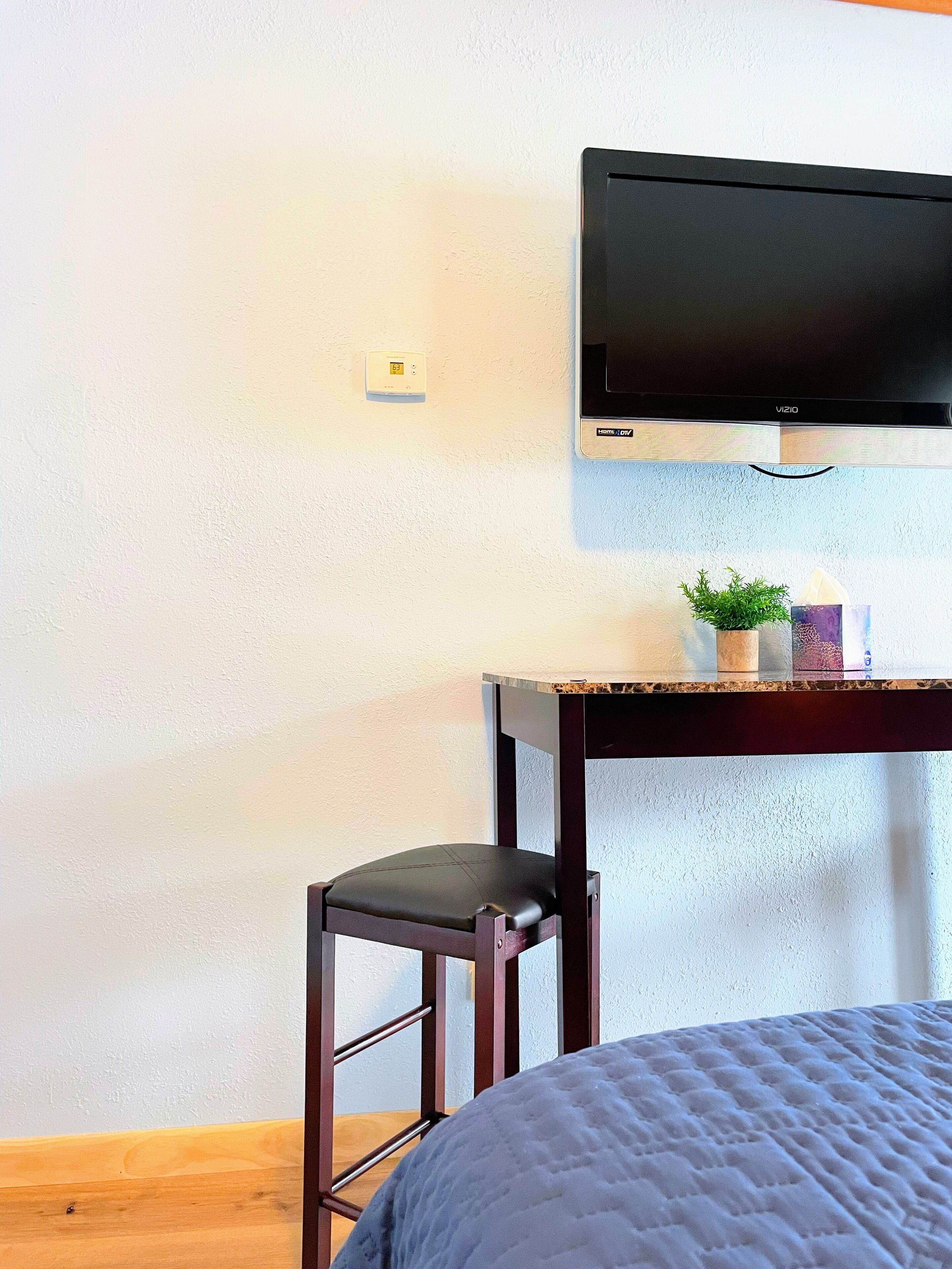 A bedroom with a bed, a desk with a chair, a television mounted on the wall, and a thermostat.