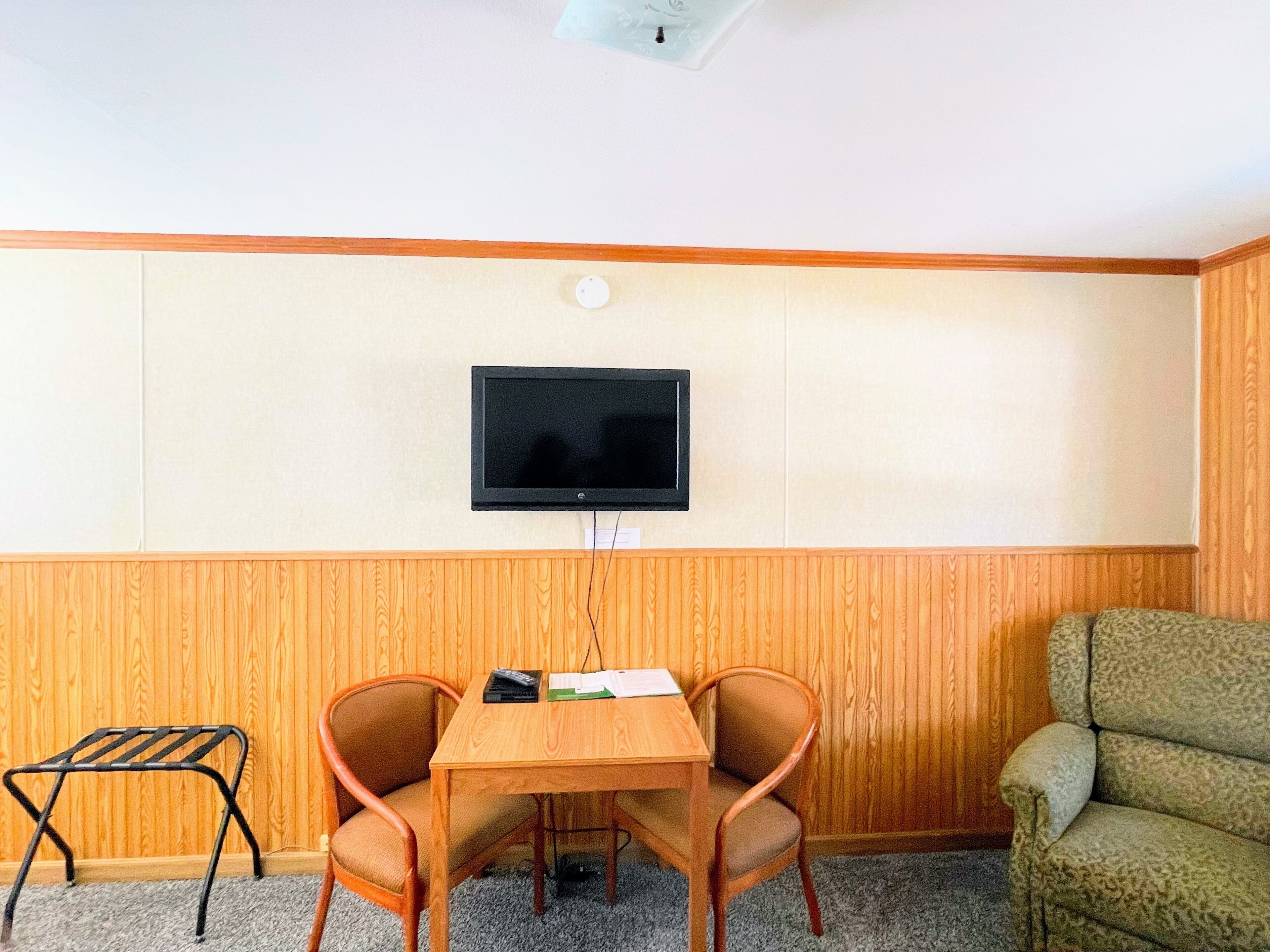 Ett rum med en väggpanel av trä, en platt-TV som hänger ovanför ett träbord med två stolar, och en grön soffa.