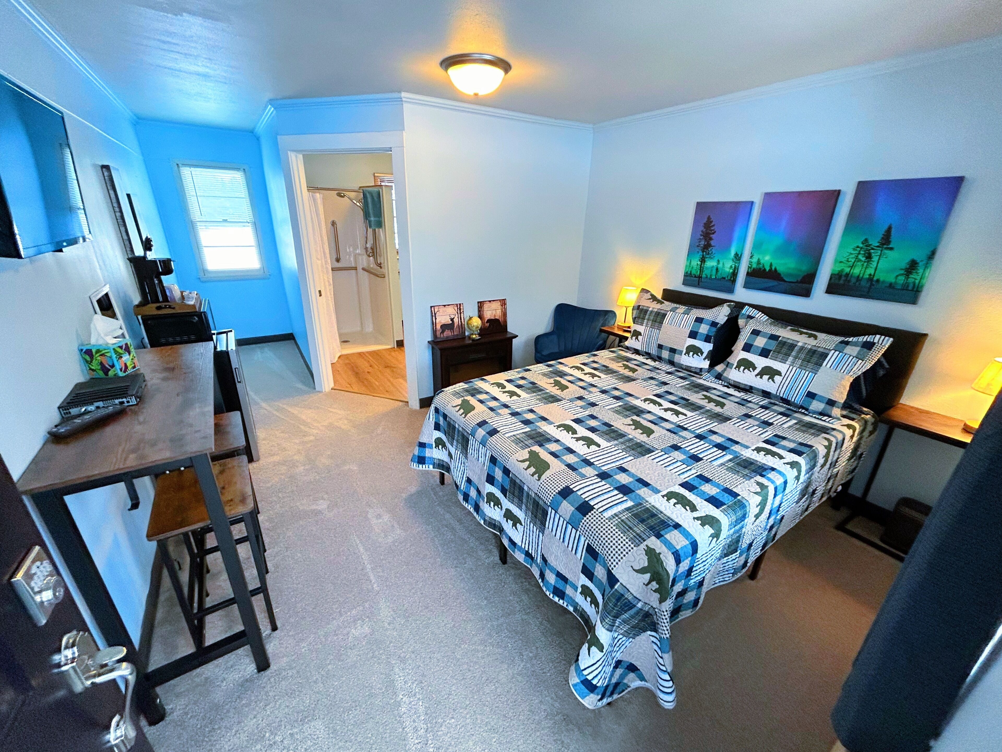 A bedroom with a large bed, a desk, and a chair. There are three paintings on the wall, a lamp on the desk, and a window with blinds.