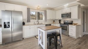 A spacious kitchen with white cabinetry, a stainless steel refrigerator, a microwave, and an island with stools. - NEW! ♦ Fun & Modern Lake Retreat ♦ Near Marina! (Pottsboro)