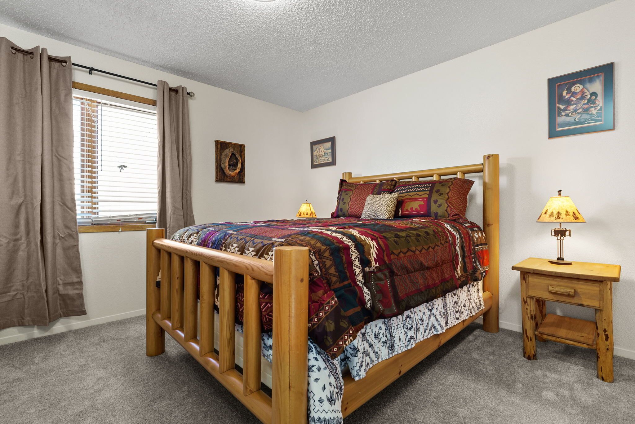 A bedroom with a wooden bed, a window with curtains, a bedside table with a lamp, and two wall-mounted pictures.