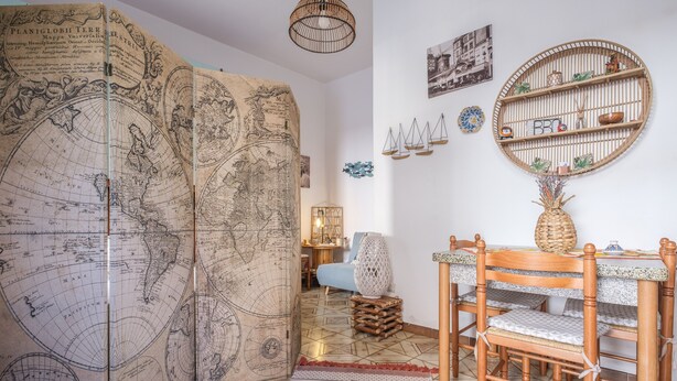A room with a large world map, a round wooden shelf with decorative items, a wooden dining table with chairs, and a patterned rug. - Holiday Home "Tyndaris" with Wi-Fi & Air Conditioning (Oliveri)