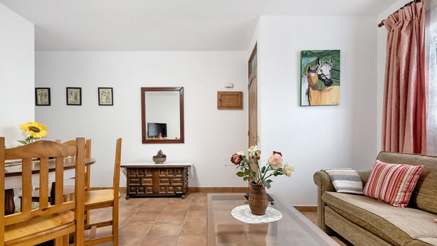 Ein Wohnzimmer mit Esstisch aus Holz, Stühlen, einer Couch, einem Couchtisch mit Blumenvase, einem Fernseher und gerahmten Kunstwerken an der Wand.