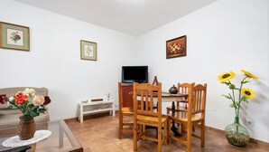 Ein Wohnzimmer mit Sofa, Couchtisch, Fernseher, Esstisch mit Stühlen und einer Vase mit Blumen.