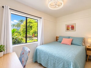 A bedroom with a bed, a window with curtains, a ceiling fan, a bedside table, a lamp, and a framed picture on the wall. - Cozy 2br 1ba Apt in Waiakea Villas with work space (Hilo)