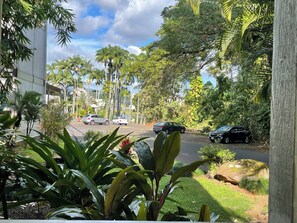 A street lined with palm trees and parked cars, with a building on the left and a grassy area on the right. - Cozy 2br 1ba Apt in Waiakea Villas with work space (Hilo)