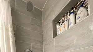 Douchegedeelte met grijze tegels, een houten plank met toiletartikelen, en een douchegordijn.