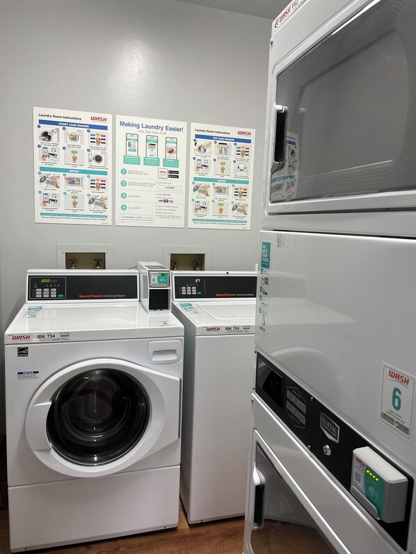 A laundry facility with multiple washers and dryers, instructional posters on the wall, and a digital display on the washers. - Cozy 2br 1ba Apt in Waiakea Villas with work space  (Hilo)