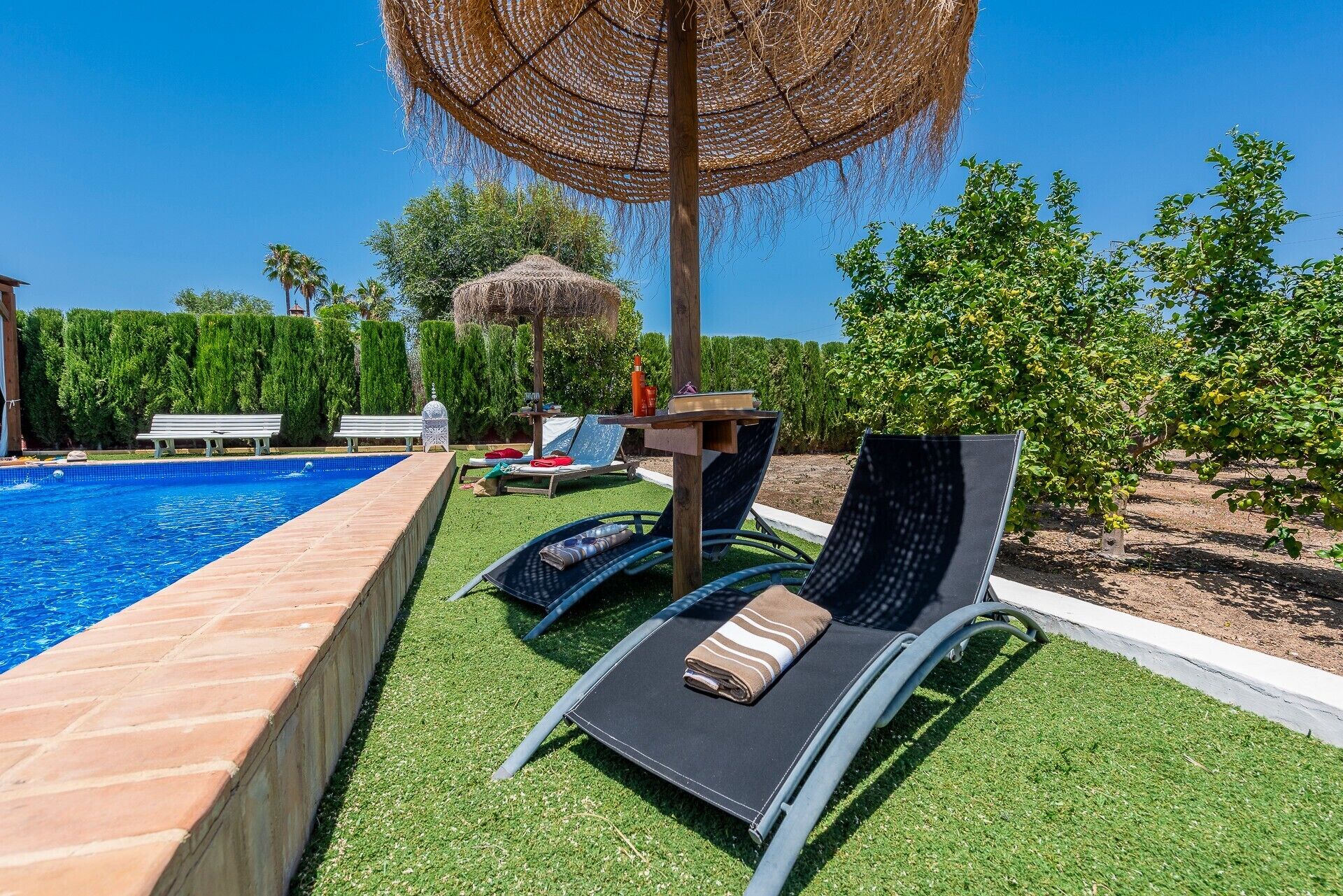 Zona junto a la piscina con sillones de descanso, una mesa y una pérgola.