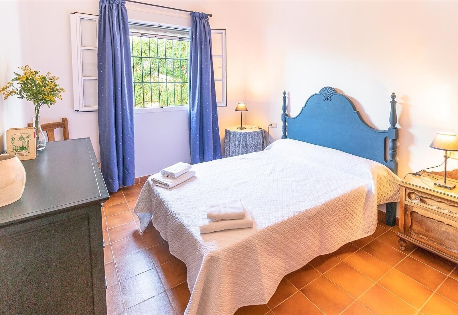 Un dormitorio con cama, mesita de noche, lámpara, ventana con cortinas, una silla y una pequeña mesa con un florero.