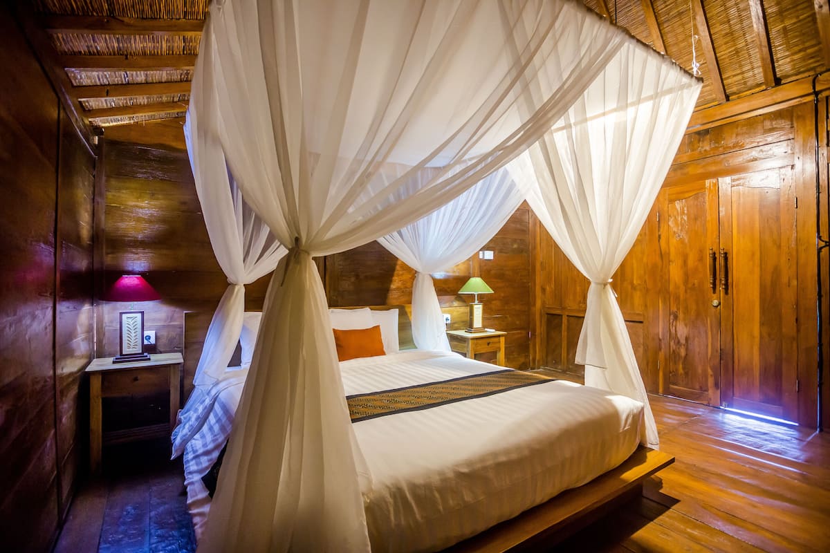 Een slaapkamer in een houten chalet met een hemelbed, nachtkastje, lamp en een houten deur.